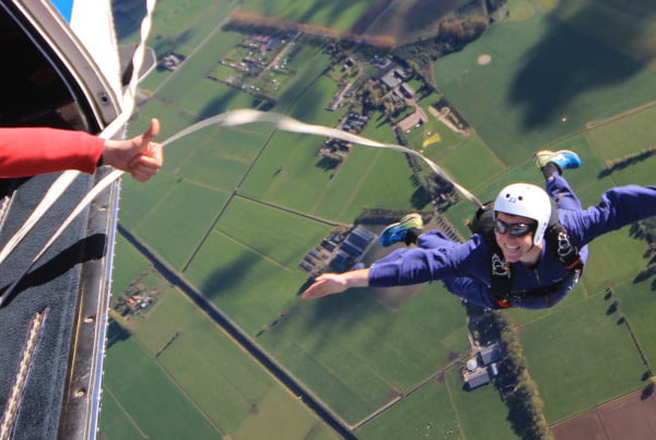 een parachutist springt uit een vliegtuig aan een static line en kijkt lachend omhoog naar de jumpmaster, die zijn duim opsteekt